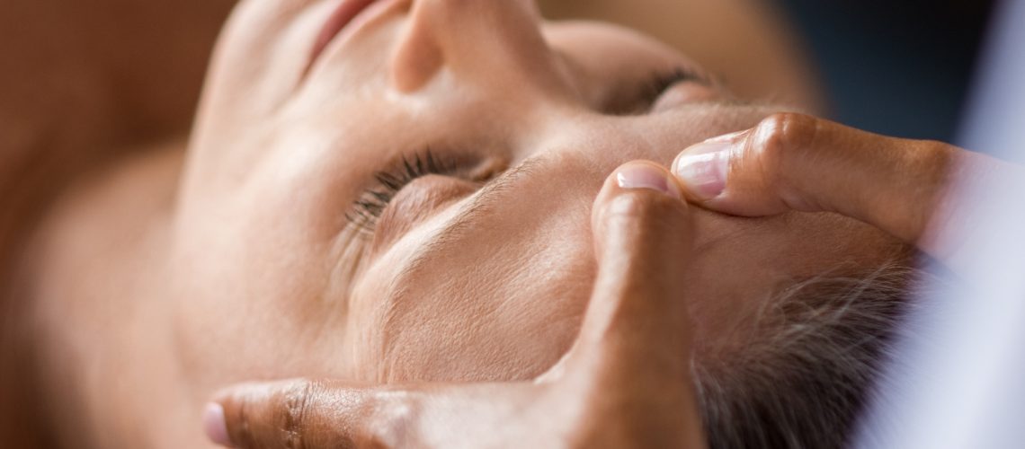 Closeup face of mature woman having facial massage at spa. Senior woman lying with closed eyes at spa while a massage therapist doing anti-aging treatment. Masseur doing head massage at wellness center.